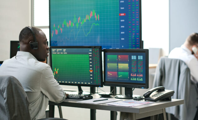 African American and Caucasian men in formalwear sitting in front of computer monitors analyzing currency stats