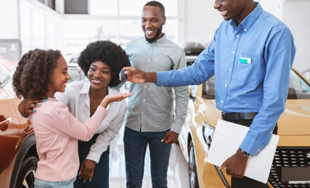 Positive auto salesman giving car key to cute black girl and her parents at dealership store. Cheerful African American family shopping for new vehicle at automotive showroom