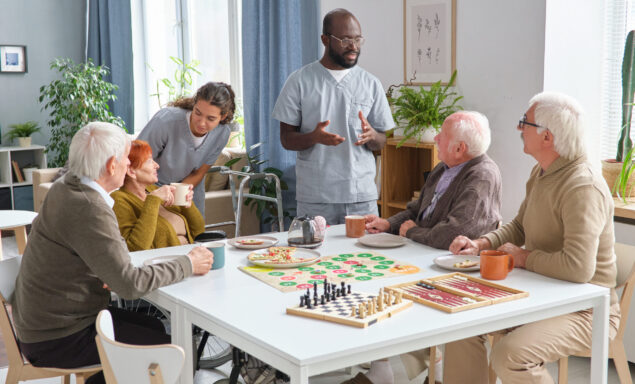 Group of senior people playing board game at the table and talking to medical staff in the room of nursing home